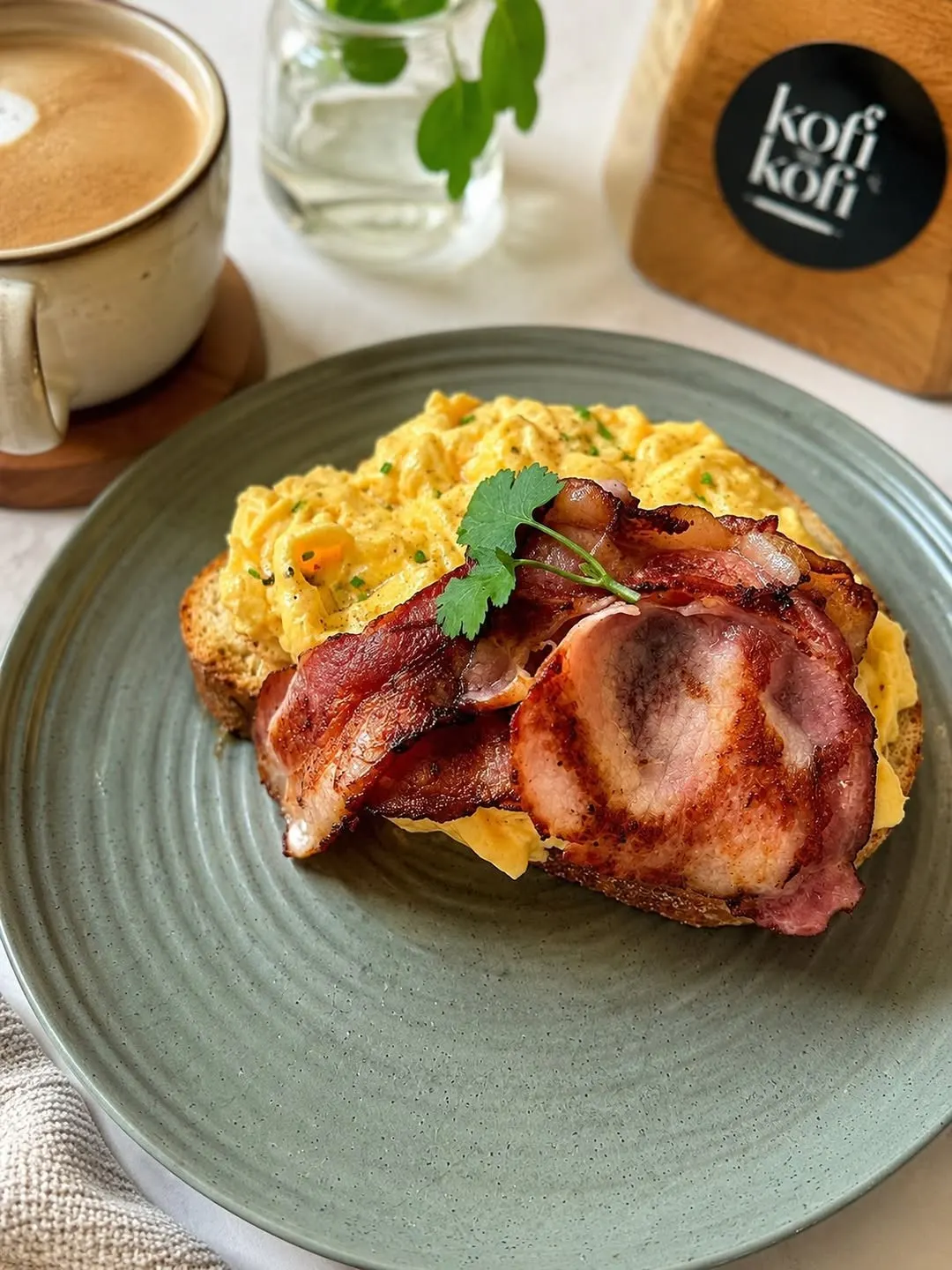 Scrambled eggs and crispy bacon on toast next to a flat white at Köfi Köfi