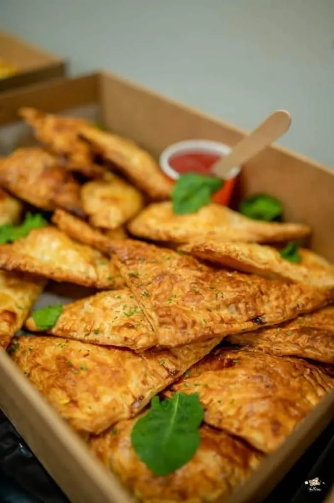 A catering box of golden triangular pastries with tomato sauce and fresh herbs